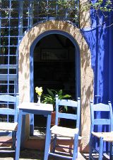 Blaue Stühle und ein Tisch vor einer blau-gelben Häuserfront mit Fenstern und offener Tür.