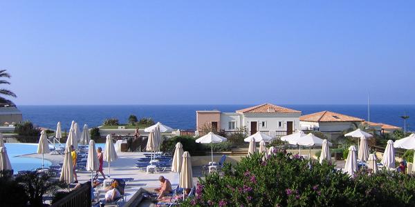 Sicht auf das Club-Hotel-Gelände mit Pool, Sonnenschirmen, weißen Häusern und blauem Meer und Himmel im Hintergrund.